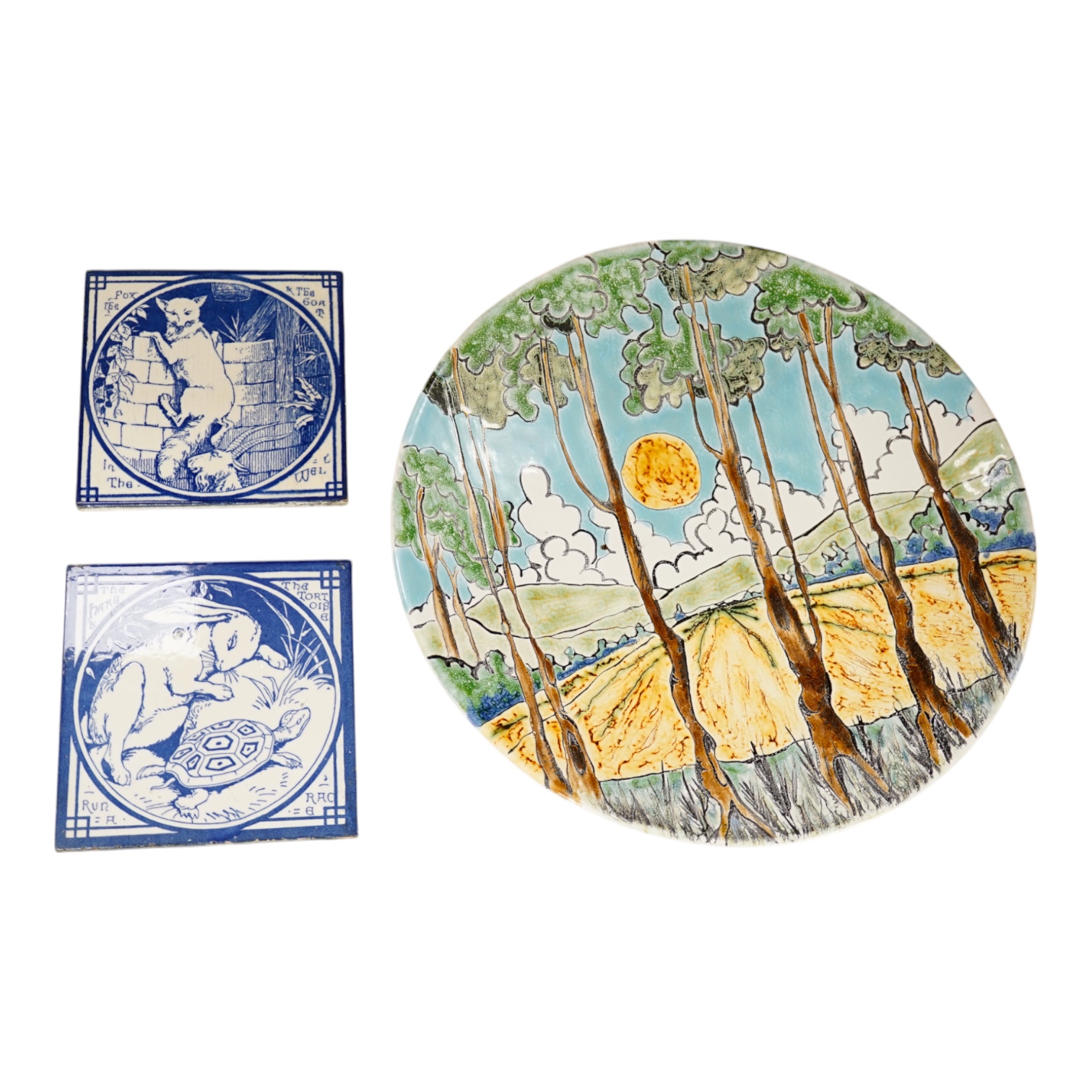 Two Minton pottery Arts and Crafts fireplace tiles, printed in blue and white, with scenes from Aesops fables - ‘’The Fox and the Goat in the Well’’ and ‘’The Hare and the Tortoise’’, together with a Poole pottery dish.